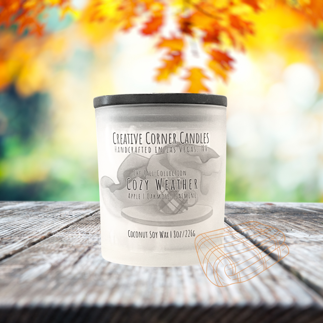 Cozy Weather fall candle with apple, oakmoss, and jasmine scent in coconut soy wax, hand-drawn label illustration of a coffee mug on wooden stand with blankets behind.