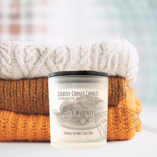 Cozy Weather fall candle with apple, oakmoss, and jasmine scent in coconut soy wax, hand-drawn label illustration of a coffee mug on wooden stand with blankets behind.