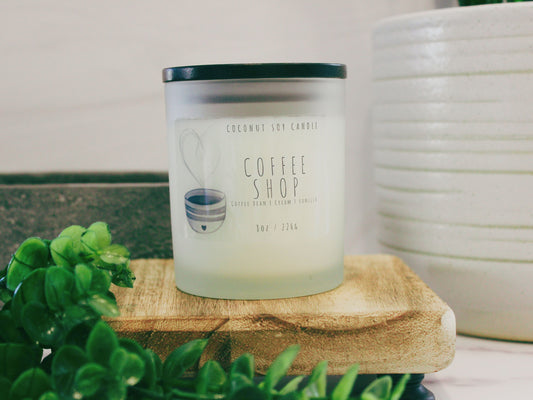 An inviting Coconut Soy Candle named 'Coffee Shop.' The candle is in a stylish glass container, emanating a warm glow. The label features a coffee cup illustration, capturing the essence of a cozy coffee shop. Perfect for creating a comforting atmosphere with rich coffee notes. Ideal for home decor and relaxation. Hand-poured and crafted with premium coconut soy wax for a clean burn. Elevate your space with this aromatic coffee-inspired candle